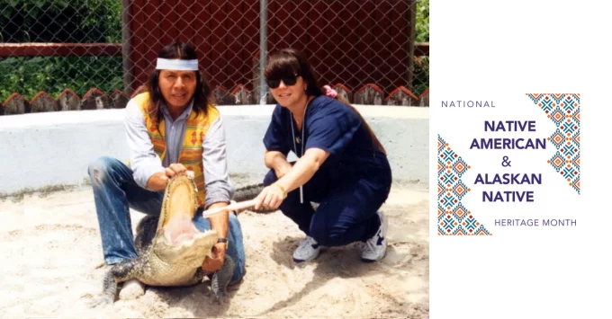 Becky in blue scrubs crouching next to a Miccosukee who is hold open an alligator mouth