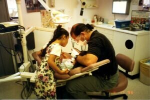 Seated dentist leans over a young patient, with patient's sibling next to him.
