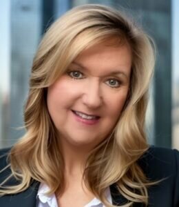 Headshot of Linda Rhoades - blond layered hair past her shoulders with a navy blue suit jacket and white blouse