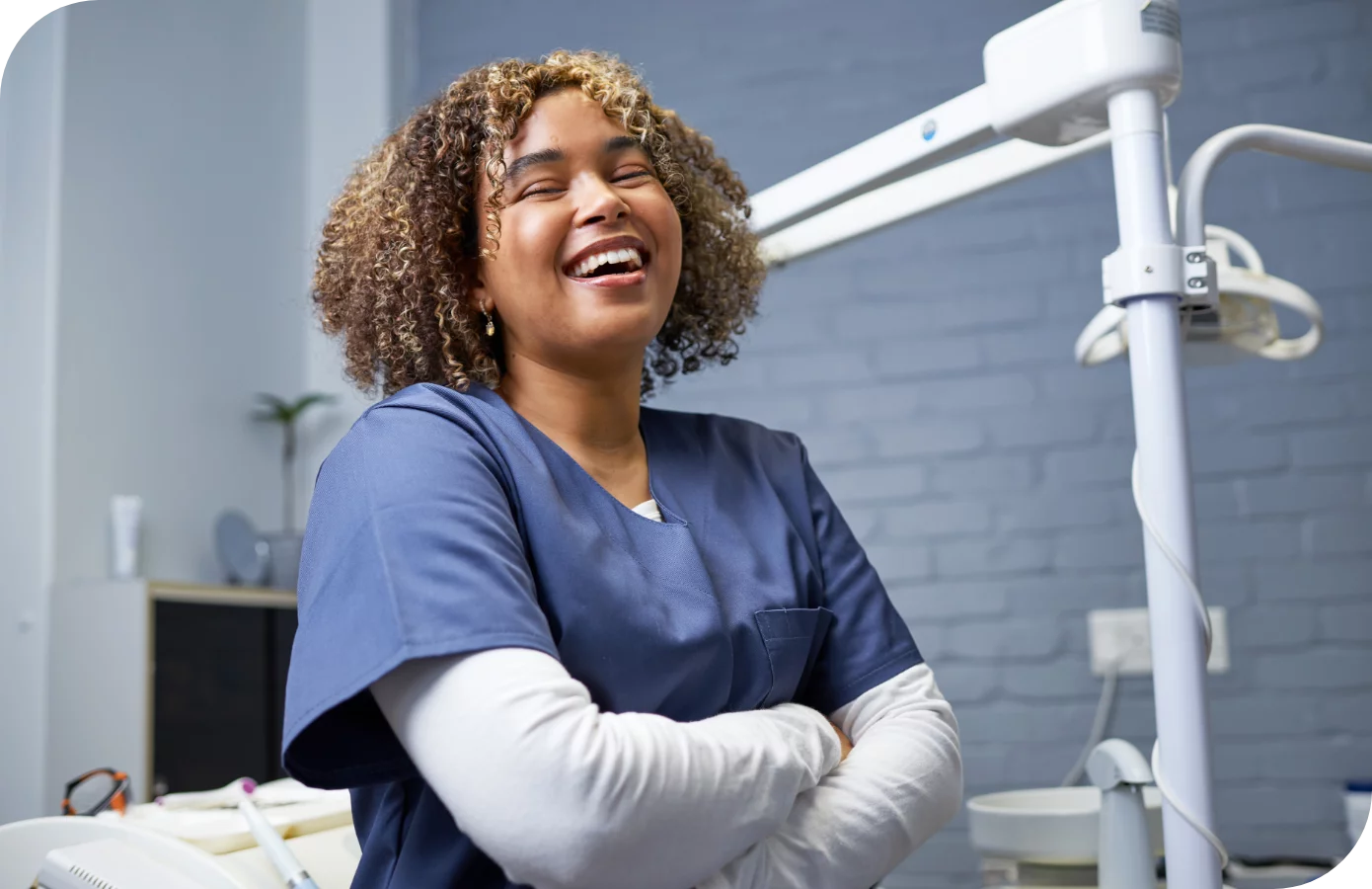 Dental hygienist with tight curls in blue scrubs with white sleeved crossed arms laughing and smiling in operatory with white brick wall