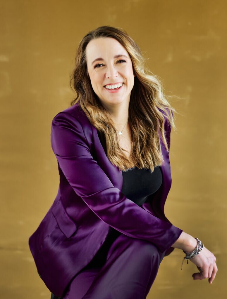 Erin Haley-Hitz in a purple silk suit sitting cross-legged on a chair with a golden background