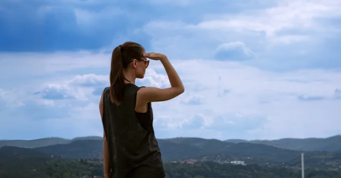 Woman in tank top standing on a hill overlooking the wide horizon landscape and shielding her eyes with her right hand