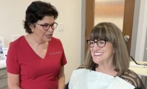 Hygienist in red scrubs sits alongside a patient in the dental operatory and talks with her