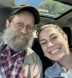 Main with cap and plaid shirt smiles in a selfie with his grown daughter in a car