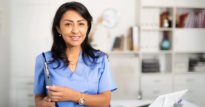 Hygienist in scrubs holding patient chart with computer and desk in background