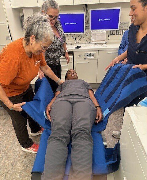 A woman in grey scrubs in a prone position is attended by three women getting ready to move her.