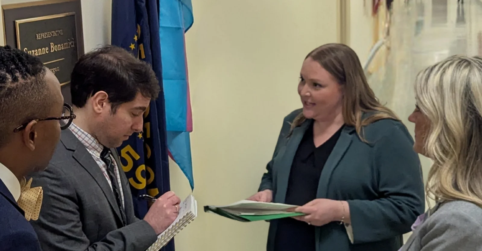 Jessica August stands holding papers in a hallway while speaking to a congressional staffer who is taking notes
