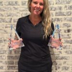 Hali Householder in black scrubs stands in front of a whitewashed brick wall holding two glass triangle-shaped ADHA awards.