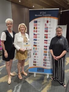 Tammy L. Filipiak and Kelli Swanson Jaecks stand left of display sign of flags from IFDH member countries and IFDH President Jill Rethman stands to the right of it, all smiling at the camera.