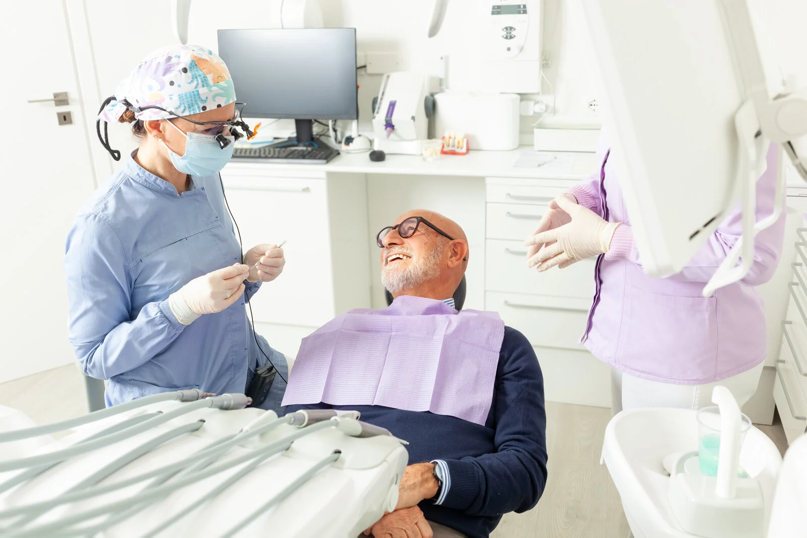 Dentist wearing magnifying glasses preparing tools for examining a senior man sitting in a dental chair, while a nurse is assisting her