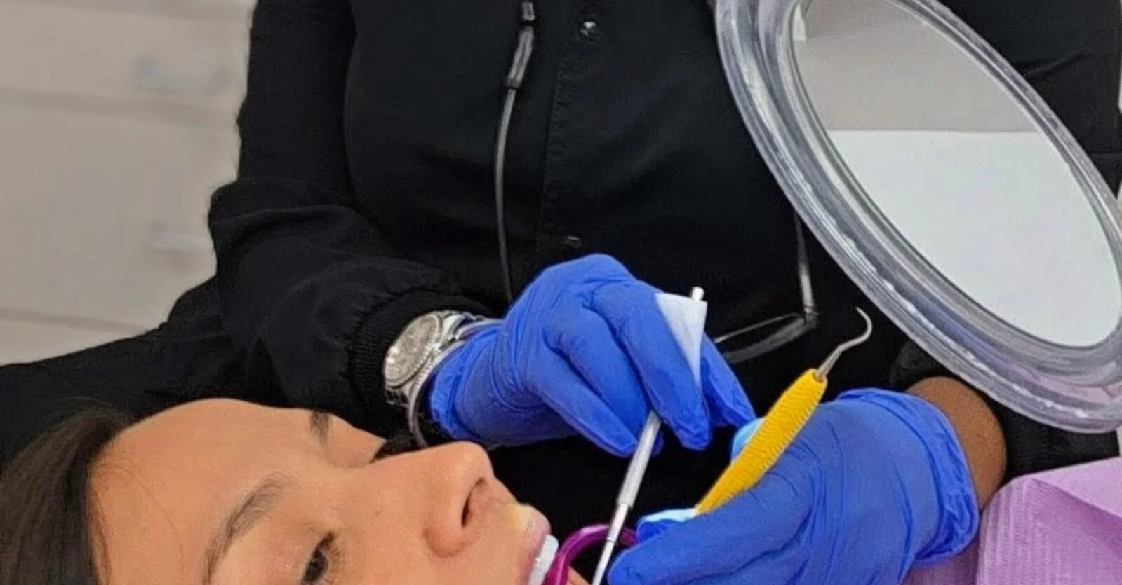A dental hygienist in black scrubs and blue gloves uses a periodontal probe on a reclined patient, who watches in a large hand mirror.