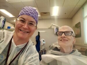 Christina Emmert, in a purple ADHA-patterned surgical cap, glasses and a white disposable gown, smiles beside a patient seated in a dental chair in an operator