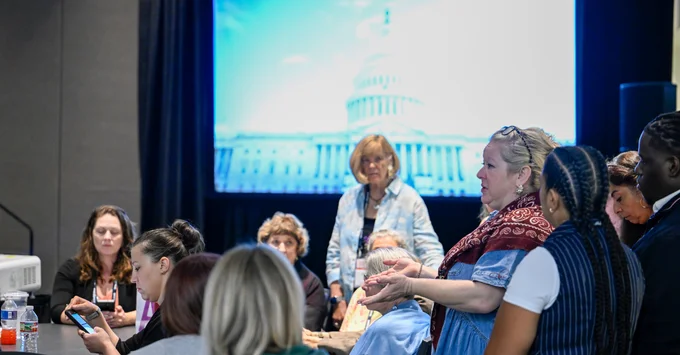 Attendees seated around tables in a meeting room while one woman gestures as she speaks and another stands behind them, with a projected image of the U.S. Capitol on the screen behind.