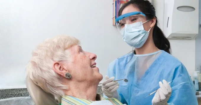 A dental hygienist in a face mask, protective eyewear, and blue gown leans over an older woman reclined in a dental chair, holding a mirror and explorer while the patient smiles up at her.