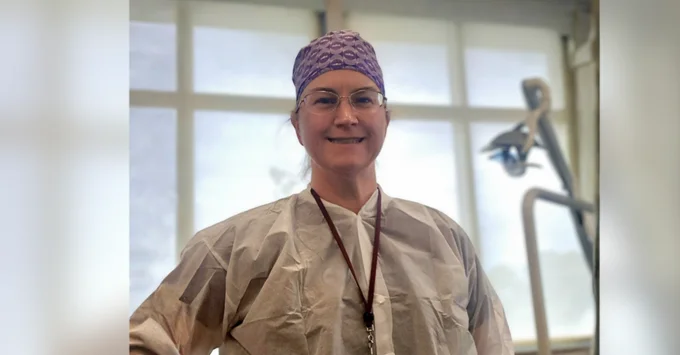 Christina Emmert, wearing a purple patterned surgical cap, glasses, and a white disposable lab coat with a maroon lanyard, smiles at the camera in a dental operatory with an operatory light and chair behind her.