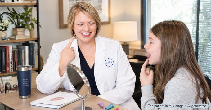 Joy Lantz in a white coat with an OFS logo smiles and points to her mouth while a young girl across the table copies her, tongue raised, during an orofacial myology session. A hand mirror sits between them. The patient in this image is AI-generated.
