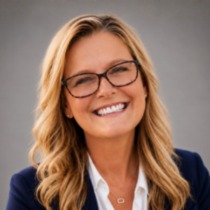 Headshot of Laureen Brady, a smiling woman with long, wavy blonde hair and tortoiseshell glasses, wearing a navy blazer over a white collared shirt and a thin gold necklace, against a gray background.